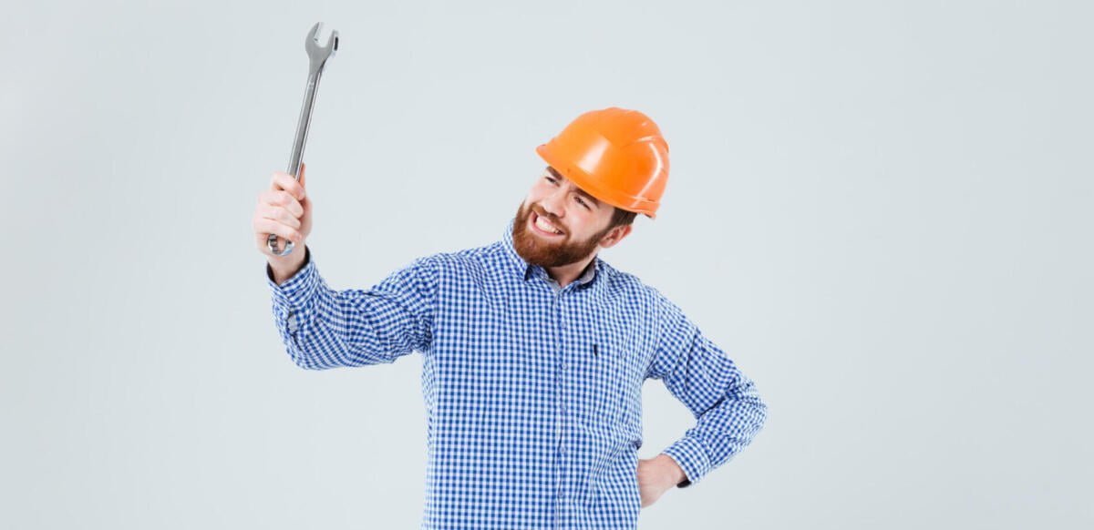 Cheerful bearded man in helmet holding and looking at wrench